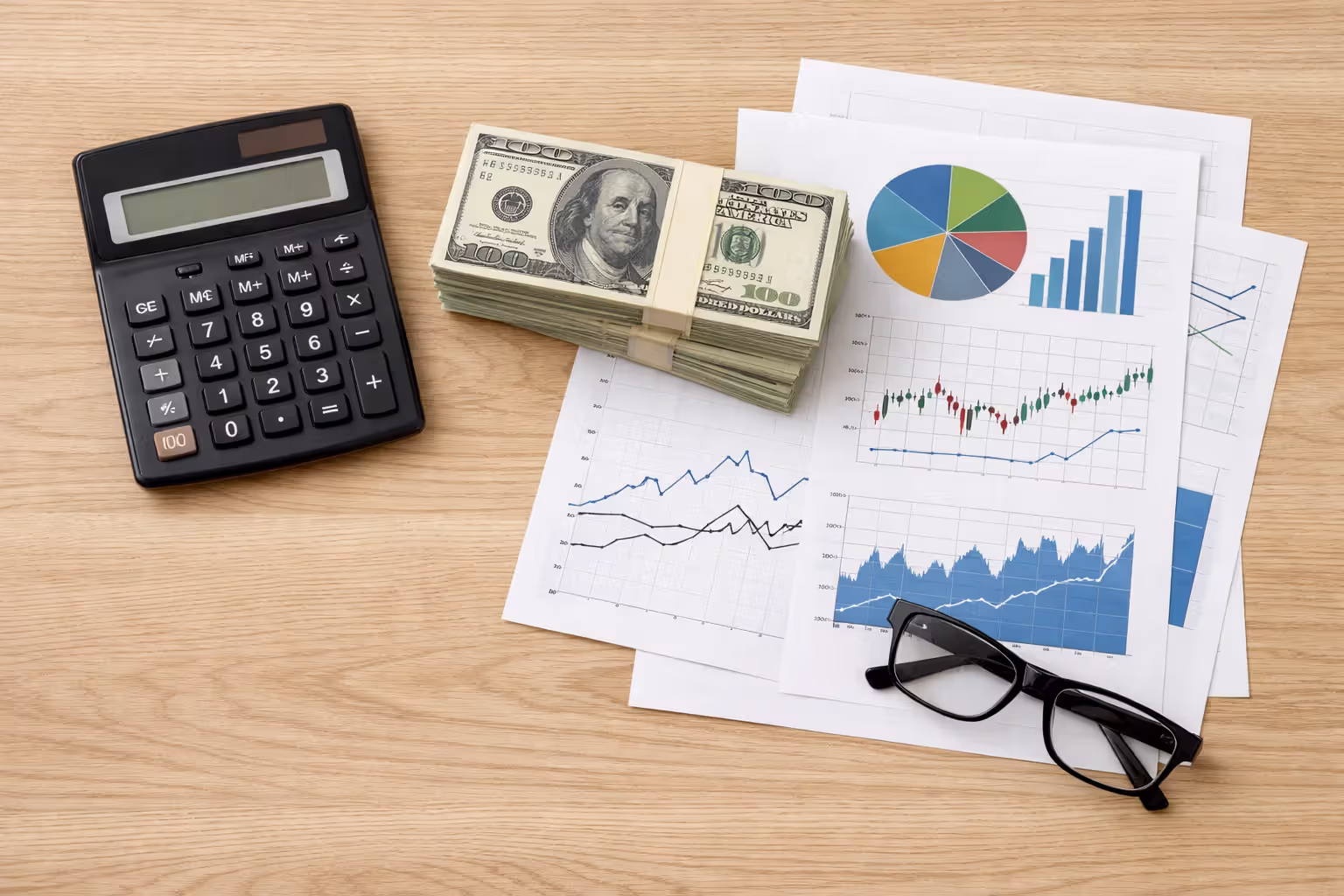 Calculator, stack of dollar bills, financial charts on printed sheets, and eyeglasses on a wooden desk viewed from above